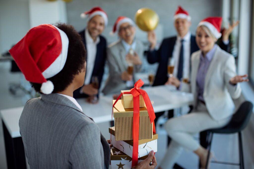 Eine Person mit Weihnachtsmütze übergibt gestapelte Geschenke an ein Team von Kollegen in festlicher Atmosphäre – ideale Szene für Kundengeschenke oder Mitarbeitergeschenke zu Weihnachten.