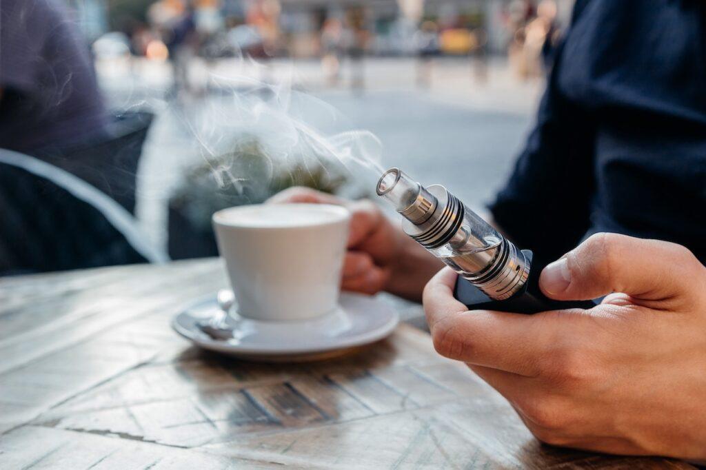 Ein Tisch in einem Café mit einer dampfenden Kaffeetasse im Vordergrund. Eine Person hält eine silberne E-Zigarette (Vape) in der Hand, aus der Dampf aufsteigt. Der Hintergrund zeigt eine leicht verschwommene urbane Szene. Das Bild vermittelt moderne Genussmomente im Café-Alltag.