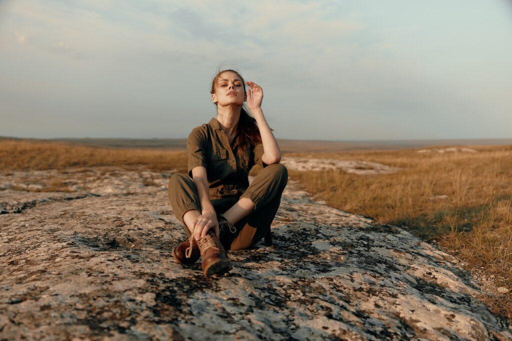 Eine junge Frau sitzt entspannt im Schneidersitz auf einem flachen Felsen inmitten einer weiten, grasbewachsenen Landschaft. Sie trägt einen olivgrünen Overall und robuste Wanderstiefel. Mit geschlossenen Augen genießt sie den Moment in der warmen Abendsonne, die die Szene in weiches Licht taucht. Die Atmosphäre strahlt Ruhe, Freiheit und Naturverbundenheit aus.